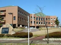 建物を屋外にて左斜め前から撮影した写真。建物は3階建てかつオレンジ色の壁でできており、建物の前には石碑がある。