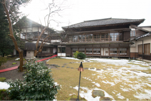 手前にうっすら雪が残った広い芝生の庭園があり、奥に木造2階建ての旧五十嵐家住宅主屋がある写真