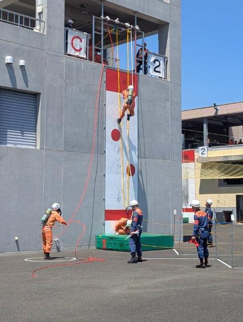 屋外の大会会場で、出場した救助隊員の一人が約8メートルの塔の壁を登り、それを補助する隊員と審査員が見守る様子