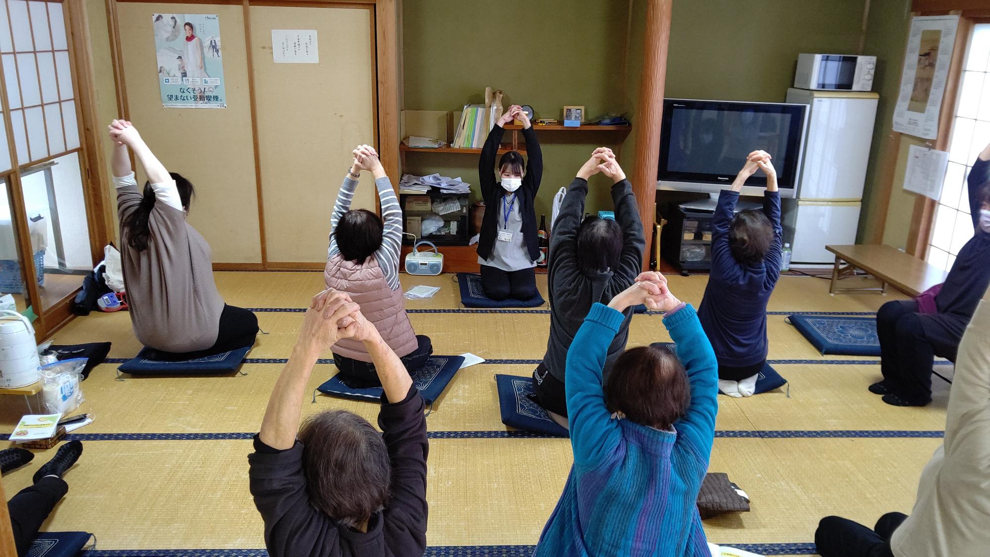 和室で参加者の皆さんが立って後ろに両手を組み片足を後ろに伸ばす体操をしている写真