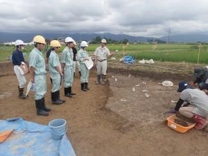 ヘルメットを被り作業服を着て長靴をはいて見学している4人の高校生と発掘作業をしている作業員の写真