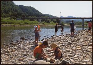 河川敷で石を積んで遊んでいる2人の子ども、その後ろに虫取り網をもって立っている子ども、川の中に入っていく子供たちの写真