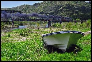 煙を上げて走るSLと川岸に置かれた1艘の舟を写した写真
