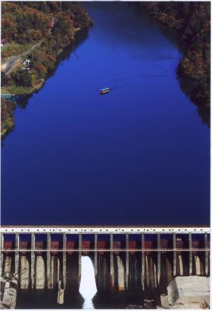 真ん中に一艘の船が浮かぶ、濃く深い青色の川と放水しているダムの写真