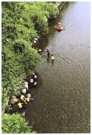 川の浅瀬で黄色いライフジャケットを着て魚取りをしている子供たち、傍で網を構えている男性、赤いカヌーの写真