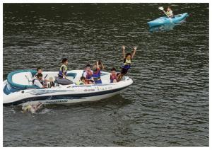 川でボートに乗っている8人の子供のうち1人の男の子がボートの先端から両手を挙げて川に飛び込もうとしている写真