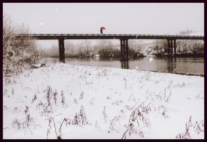 一面雪で真っ白の川岸と川にかかっている橋の上を赤い傘をさして歩いている人物が映っている写真
