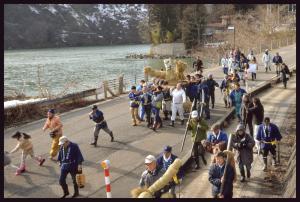 大きな鍾馗様の藁人形を担いで川沿いの道を練り歩いている祭りの参加者たちの写真