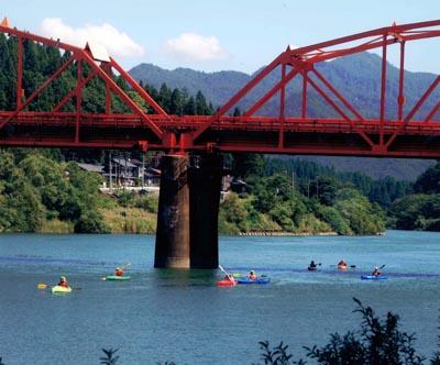 川にかかる大きな赤い色の鉄橋と黄色、緑、青、赤のカヌーに乗って遊んでいる人々の写真
