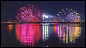 橋の向こうで打ちあがったピンクと黄色の花火が水面に写っている川の写真