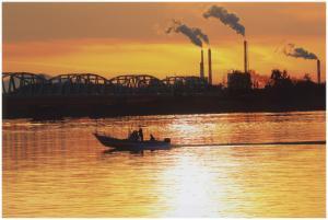 オレンジ色の夕焼け空と川、工場の煙突から煙が出ており、川の真ん中に船のシルエットが写っている写真