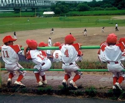 赤と白のユニフォームを着て緑色の柵に座って野球の試合を眺めている子供たちの写真