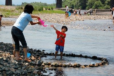 ピンク色のじょうろを持った母親と小さな男の子が河原で水遊びをしている写真