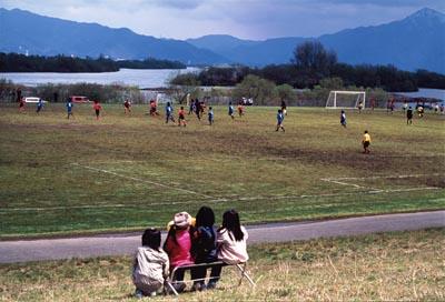 河川敷で行われているサッカーの試合をベンチに座って眺めている4人の女の子の写真