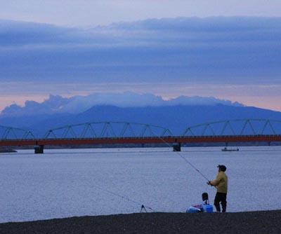 空に薄く雲がかかり、山の向こうが少し明るくなっていて早朝に釣りをしている釣り人が写っている写真