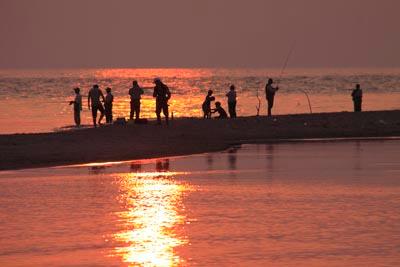 夕焼け空が反射して水面がオレンジ色に輝いている川と堤防に立って釣りをしている釣り人たちの写真