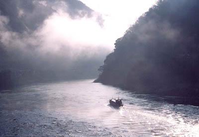 薄い雲の隙間から光がさしている川を一艘の船が進んでいる写真