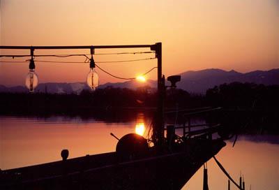 朝日が昇り、空と川の水面がオレンジ色に輝いて、手前には船のシルエットが写っている写真