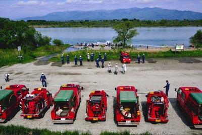 川岸の広場に横一列に整列して並んでいる消防車と川で演習をしている消防隊員の写真