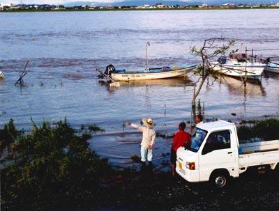 増水した川に浮かぶ2艘のボートと軽トラックの横で川を眺めている3人の男性の写真