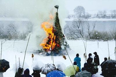 雪が降りしきる中燃えている木と傘をさしてどんど焼きを眺めている人々の写真