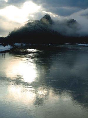 太陽の光が水面に反射している川と雲がかかった山の写真