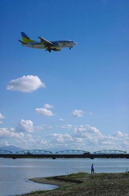 青い空を飛んでいく飛行機と川岸に立っている人物の写真