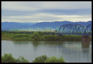 白い雲がかかった空、遠くまで連なる山脈、広々とした大きな川にかかっている鉄橋の写真