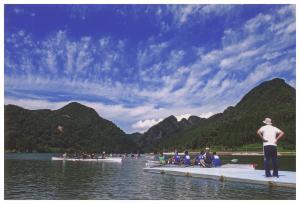 青い空と白い雲の下には山々が連なっており、川では人々がボートに乗っている写真
