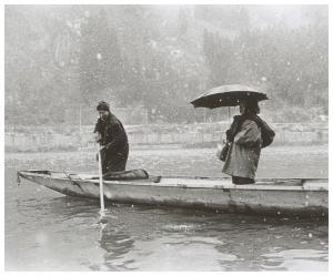 雪が降っている中、船をこいでいる人物と船に乗って傘をさしている人物が写っている白黒写真