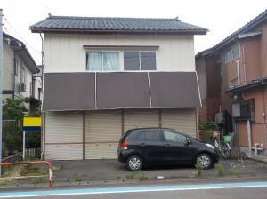 一階にはシャッターがついている2階建ての物件と家の前に停まっている黒い軽自動車の写真