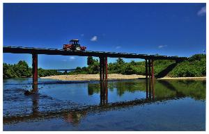 のどかな川に掛かった鉄橋を、一台のトラクターが通っている写真