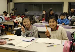 2人の女の子が机に向かい笑顔で鉛筆を持っており、その後ろにはたくさんの子供達が勉強をしている写真