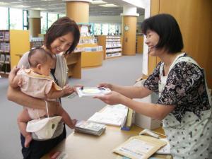 本棚がたくさんある受付で新生児を抱っこしたお母さんに絵本を渡している女性の写真
