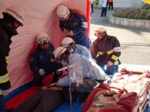 担架の横に仰向けに寝ている男性と男性を救護している5人の救急隊員の写真