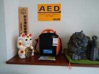 壁に貼られたAED設置のステッカーとその下にある黒とオレンジ色のAEDの写真