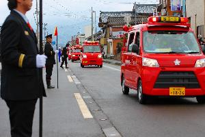 道路を一列に並んで行進する消防団の車の写真